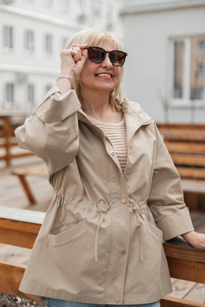 Beautiful cheerful elderly woman model in a beige fashionable windbreaker and sweater puts on sunglasses and walks on a spring day in the city - Foto, Imagem