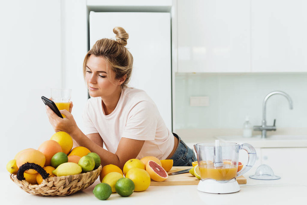 Young woman drinking freshly squeezed homemade orange juice and using smartphone in modern white kitchen - Foto, afbeelding