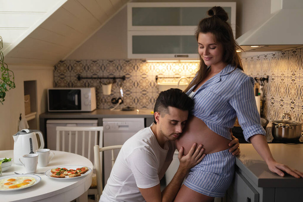 Young and sensual couple waiting for a baby. Husband and his pregnant wife during breakfast time in cozy kitchen. - Photo, Image