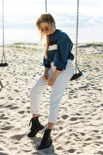 Young pretty woman is sitting on the swing and smiling at the sandy beach. - 写真・画像