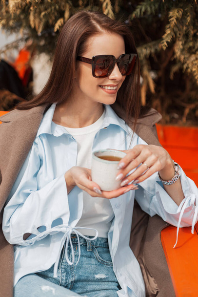 Güneş gözlüklü, şık bir ceket ve tişörtle gülümseyen genç ve mutlu bir kadın. Güneşli bir günde elinde bir fincan çay tutuyor ve dışarıda çay içiyor. - Fotoğraf, Görsel