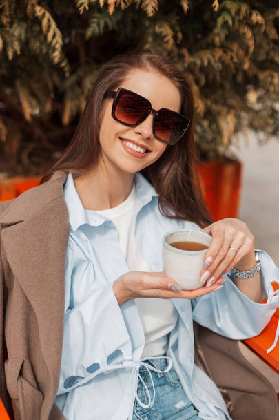 Beautiful happy woman with a cute smile with stylish sunglasses in trendy fashion clothes with coat sits in a cafe and drinks tea on a sunny warm spring day. Spring time - Zdjęcie, obraz