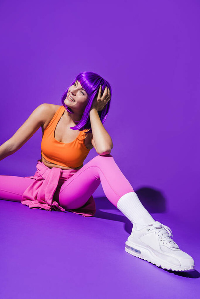 Portrait of cheerful woman wearing colorful sportswear sitting against purple background - Foto, Imagen