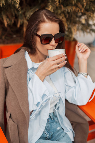 Csinos fiatal, elegáns lány napszemüveggel divatos alkalmi ruhákban, kabátban és ingben finom teát iszik, és egy kávézóban ül az utcán egy napsütéses napon. - Fotó, kép