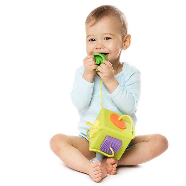 Αξιολάτρευτο χαμογελαστό μικρό αγόρι σε ένα romper κάθεται και παίζει με πλαστικό παιχνίδι, βάζοντας το στο στόμα του σε λευκό φόντο. - Φωτογραφία, εικόνα