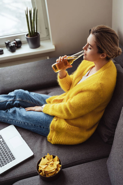 Young woman is sitting on the sofa drinking beer and eating nachos while watching movie on her laptop at home - Photo, Image