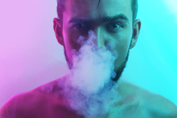 Portrait of handsome young man with wet skin in ultraviolet light smoking vape - Photo, Image