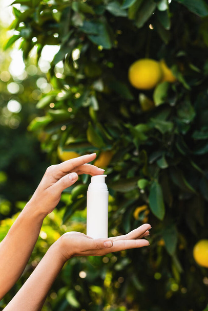 Woman holds a white bottle of skin care product in her hands on blurry lemon tree background. - Photo, Image