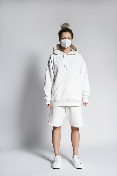 Young woman wearing white hoodie and ffp2 respirator mask against gray background - Foto, afbeelding