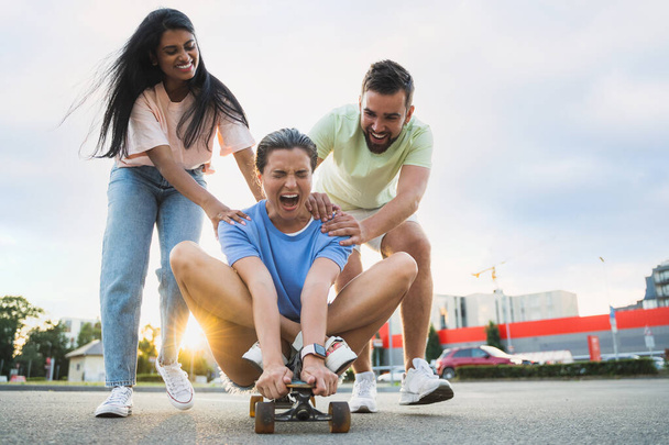 Three diverse carefree friends having fun and riding longboard on parking lot - Foto, imagen
