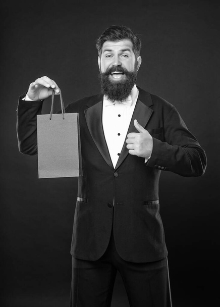amazed man in tuxedo bow tie formalwear on black background with shopping bag. birthday. copy space. thumb up. - Zdjęcie, obraz