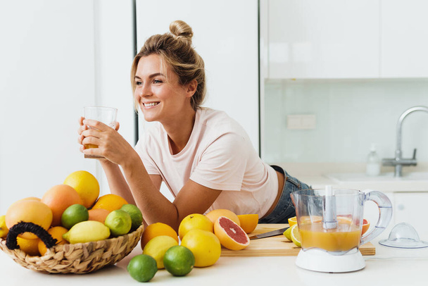 Young woman drinking freshly squeezed homemade orange juice in white kitchen - Foto, Bild