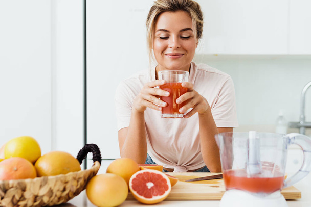 Young woman drinking freshly squeezed homemade grapefruit juice in white kitchen - Foto, afbeelding