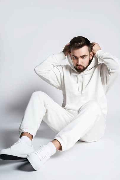 Handsome man wearing blank white hoodie and pants sitting on light gray background - Foto, imagen