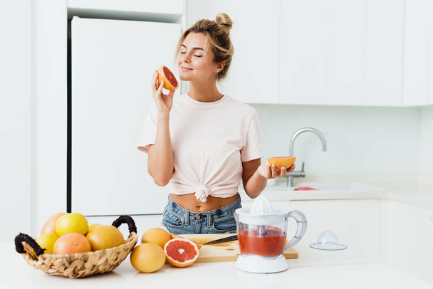 Young beautiful woman sniffing fresh grapefruit during citrus juice preparation at home - Foto, immagini