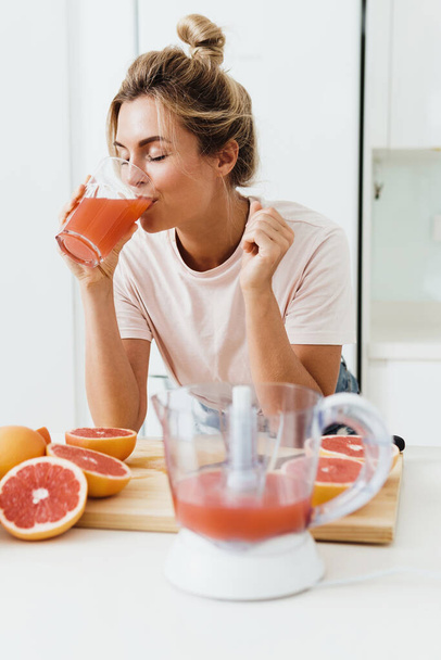 Young woman drinking freshly squeezed homemade grapefruit juice in white kitchen - Фото, зображення