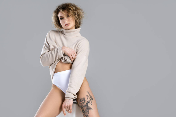 Portrait of young and sexy woman with curly blond hair wearing turtleneck jumper is posing on gray background in studio - Fotó, kép