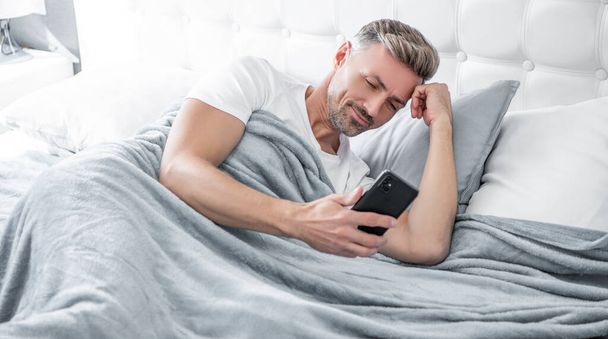 cheerful mature man in bed chatting on phone. - Фото, изображение