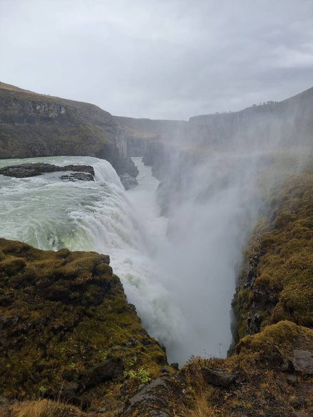 A vertical shot of the Gullfoss waterfall in Hvita, Iceland covered in steam under a cloudy sky - Fotó, kép