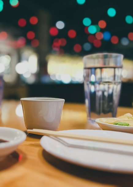A closeup shot of an interior of a restaurant, with chopsticks on a plate and a glass on a table, with bokeh lights in the background - Photo, image