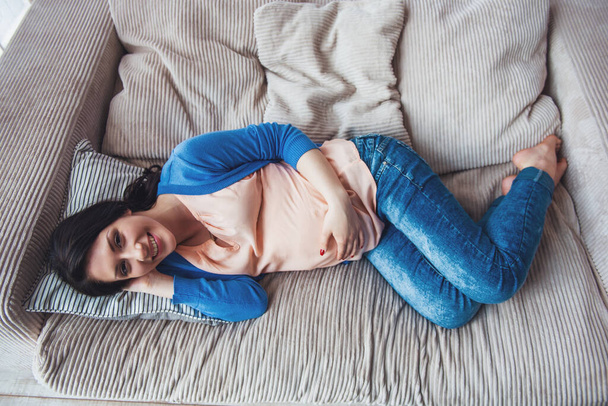 Вид сверху красивой беременной женщины, держащей одну руку на животе, смотрящей в камеру и улыбающейся, лежа дома на диване
 - Фото, изображение