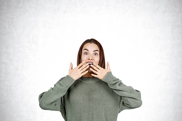Portrait of astonished young woman in green sweater with hands near open mouth standing over concrete wall background. Concept of surprise - Foto, Imagem