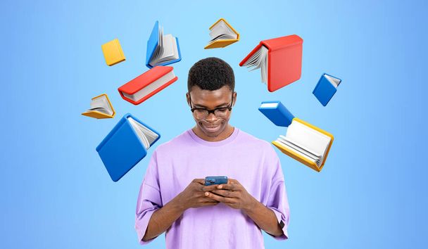 Sonriente hombre africano escribiendo en el teléfono vistiendo camiseta púrpura, flotando libros de colores sobre fondo azul. Concepto de biblioteca digital, educación, aplicación móvil y e-learning - Foto, imagen
