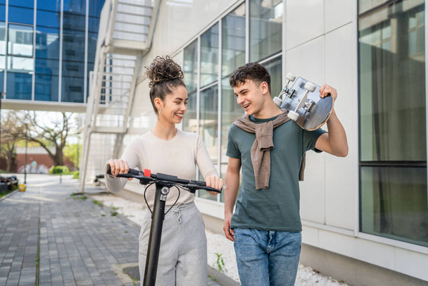 Genç adam ve kadın genç çift, beyaz erkek ve kız kardeş, mutlu bir gülümsemeyle modern binanın önünden geçiyorlar. Kaykay ve elektrikli scooter taşıyorlar. - Fotoğraf, Görsel