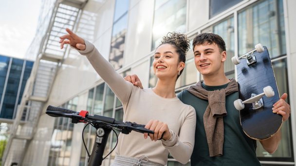 Genç adam ve kadın genç çift, beyaz erkek ve kız kardeş, mutlu bir gülümsemeyle modern binanın önünden geçiyorlar. Kaykay ve elektrikli scooter taşıyorlar. - Fotoğraf, Görsel