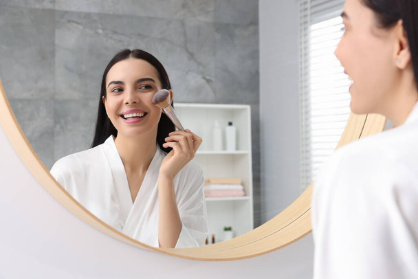 Beautiful young woman applying makeup with brush near mirror in bathroom - Fotoğraf, Görsel