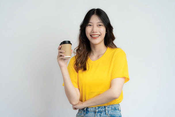 Smiling Asian girl in yellow shirt holding coffee mug standing on white background - Photo, Image