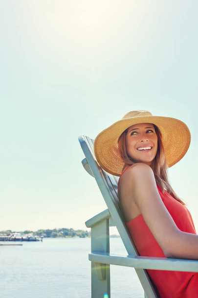 The ideal way to spend a vacation. a cheerful young woman wearing a hat while being seated on a chair next to a lake outside in the sun - Fotografie, Obrázek