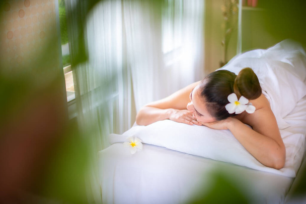 mooie aziatische vrouw liggend op een wit bed zo comfortabel dat het maakt haar in slaap vallen in spa kamer en er is een wazig groen blad in de voorkant. - Foto, afbeelding