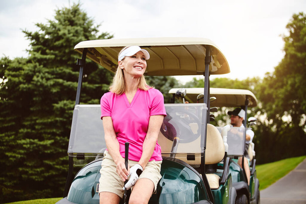 ゴルフコースは彼女の休暇のアイデアです。ゴルフ場のゴルフカートにもたれかかる魅力的な熟女 - 写真・画像