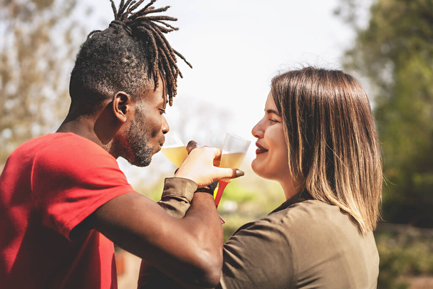 幸せな若い異人種間のカップル、アフリカ人男性と白人女性、田舎の環境で屋外でシャンパントーストを楽しんで. - 写真・画像