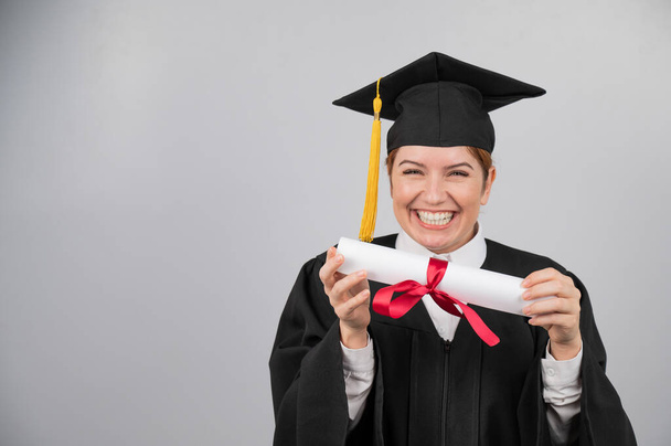 Smiling woman in graduation gown holding diploma on white background - Foto, afbeelding
