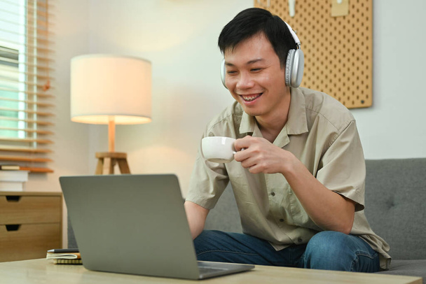 Pleasant man in wireless headphone and casual clothes drinking coffee and using laptop on couch at home. - Foto, Bild