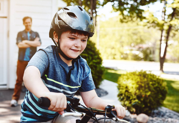 Μαθαίνοντας μια νέα ικανότητα και αγαπώντας την για μια ζωή. Ένα νεαρό αγόρι να κάνει ποδήλατο με τον πατέρα του στο βάθος. - Φωτογραφία, εικόνα