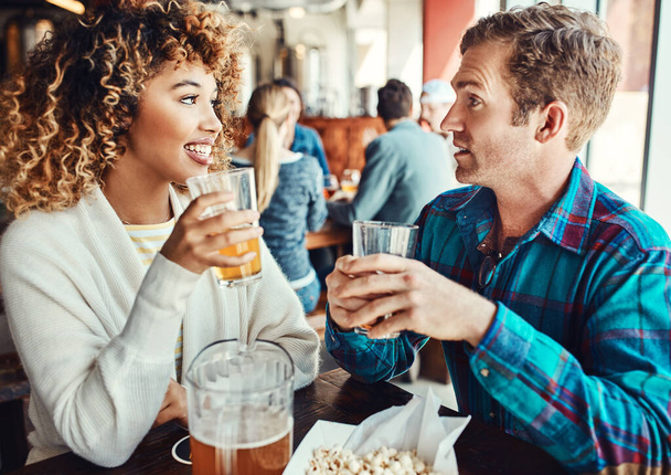 Conózcanse unos a otros por una fría. una pareja joven tomando bebidas en un bar con gente borrosa en el fondo - Foto, Imagen