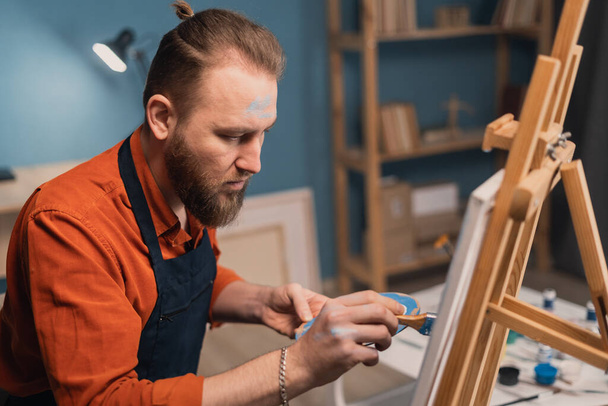 クローズアップのポートレート椅子に座っているエプロンの白人髭の男アーティストは、ブラシで油絵具で絵を描きます。キャンバスはイーゼルに設定されます。趣味と休暇のコンセプト. - 写真・画像