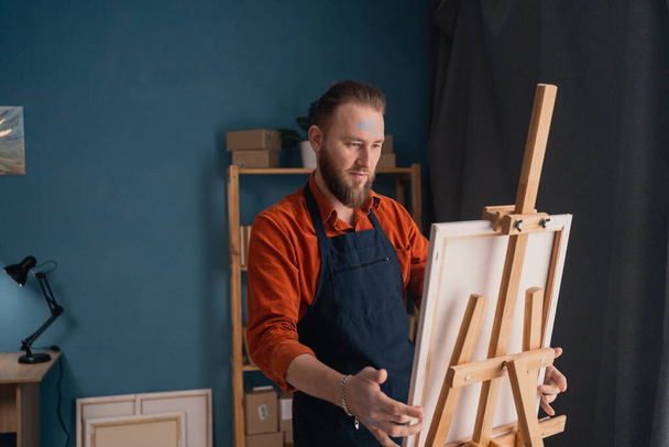 portrait caucasian bearded male artist in an apron in a home studio stands near an easel holds a canvas in his hands and looks at the picture contemptuously. hobby and vacation concept. - Fotoğraf, Görsel