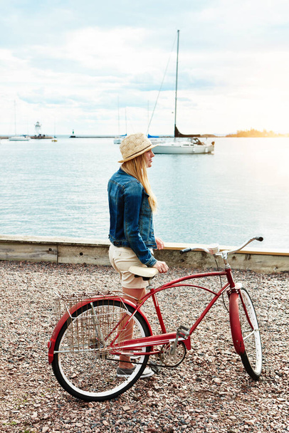 Es un hermoso día para un paseo en bicicleta. una mujer joven y tranquila con un sombrero y preparándose para montar en bicicleta mientras mira hacia el océano afuera - Foto, Imagen