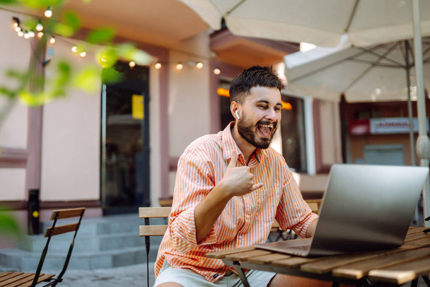 Happy male freelancer smiling while working at laptop with phone, online conference. Young man works in street cafe. Freelance business concept. Blog, education concept. - 写真・画像