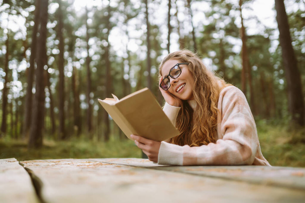 Happy young woman is sitting at table with book in beautiful forest. Beautiful woman is enjoying fresh air in forest reading book. Concept of relaxation, solitude with nature, lifestyle. - Photo, Image