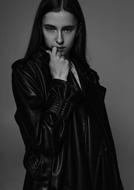 Young beautiful fashion model in long leather coat and dark jeans on dark grey. - Фото, зображення