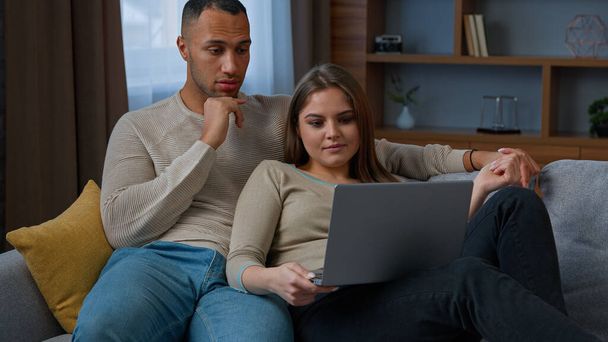 Multiracial ethnic couple homeowners shopping online use laptop hug on sofa at home diverse woman wife and man husband talk choose apartment on real estate website make order online buy internet goods - Foto, afbeelding