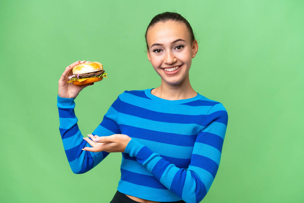 Młoda Arabka trzyma hamburgera na odosobnionym tle wyciągając ręce na bok za zaproszenie przyjść - Zdjęcie, obraz