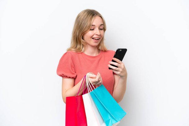Young English woman isolated on white background holding shopping bags and writing a message with her cell phone to a friend - Photo, image