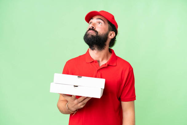 Repartidor de pizza con uniforme de trabajo recogiendo cajas de pizza sobre fondo de clave de croma aislado y mirando hacia arriba - Foto, imagen