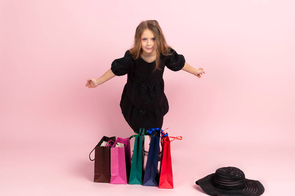 Kinder winkelen. Charmant klein meisje in zwarte stijlvolle zomerjurk en lichte roze make-up, staat tussen kleurrijke boodschappentassen. Geïsoleerd op roze achtergrond. - Foto, afbeelding